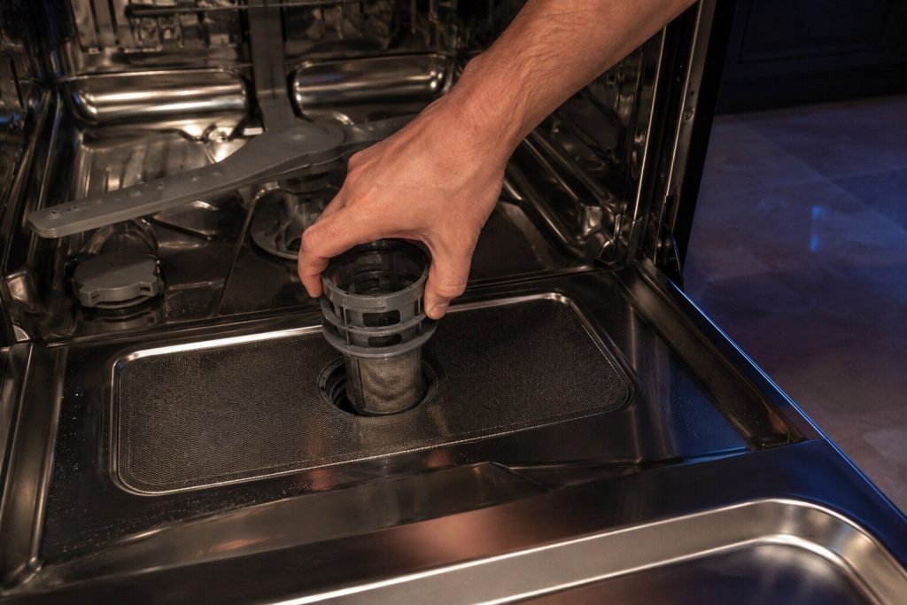 "A hand turning the cylindrical filter counter-clockwise out of the floor of a dishwasher tub, the flat mesh plate still in place beneath it"