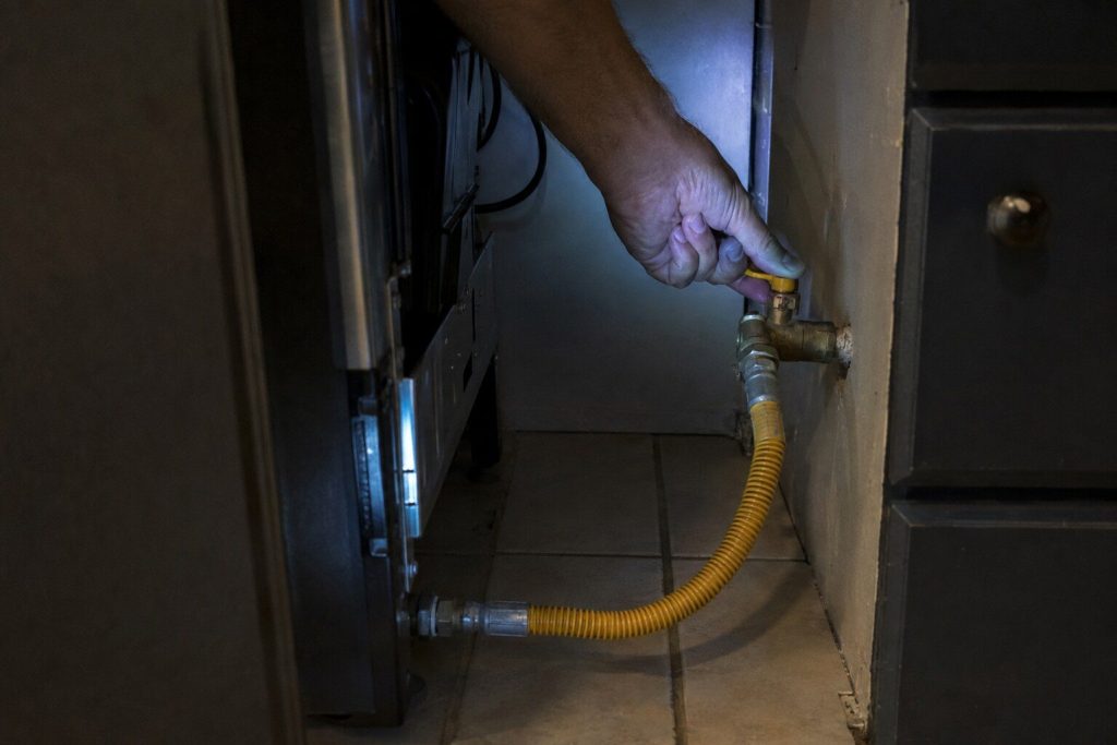 A hand closing the lever of a gas shut-off valve on the supply pipe at the wall behind a freestanding gas range that has been pulled forward onto the tile, the flexible connector looping down from the back of the range to the valve.