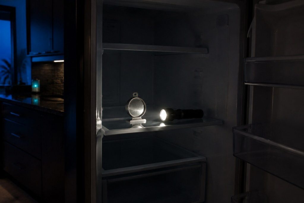"An appliance thermometer standing on a refrigerator shelf photographed by torchlight with the interior lamp off and the compartment dark, a torch lying on the shelf beside it" — storm season, measured by torch