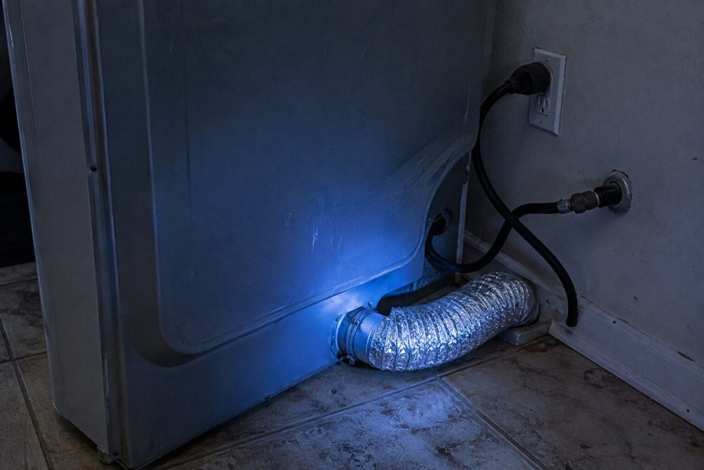 The back panel of a dryer pulled out from the wall: the round vent outlet low on the panel with a foil duct clamped to it and turning into an elbow at the baseboard, the power cord and the gas line dropping beside it.