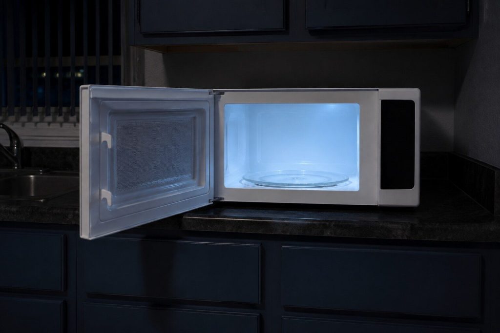 A white countertop microwave standing free on a laminate counter with its door swung fully open on an empty lit cavity and a glass turntable, the keypad column beside the door dark and unmarked.