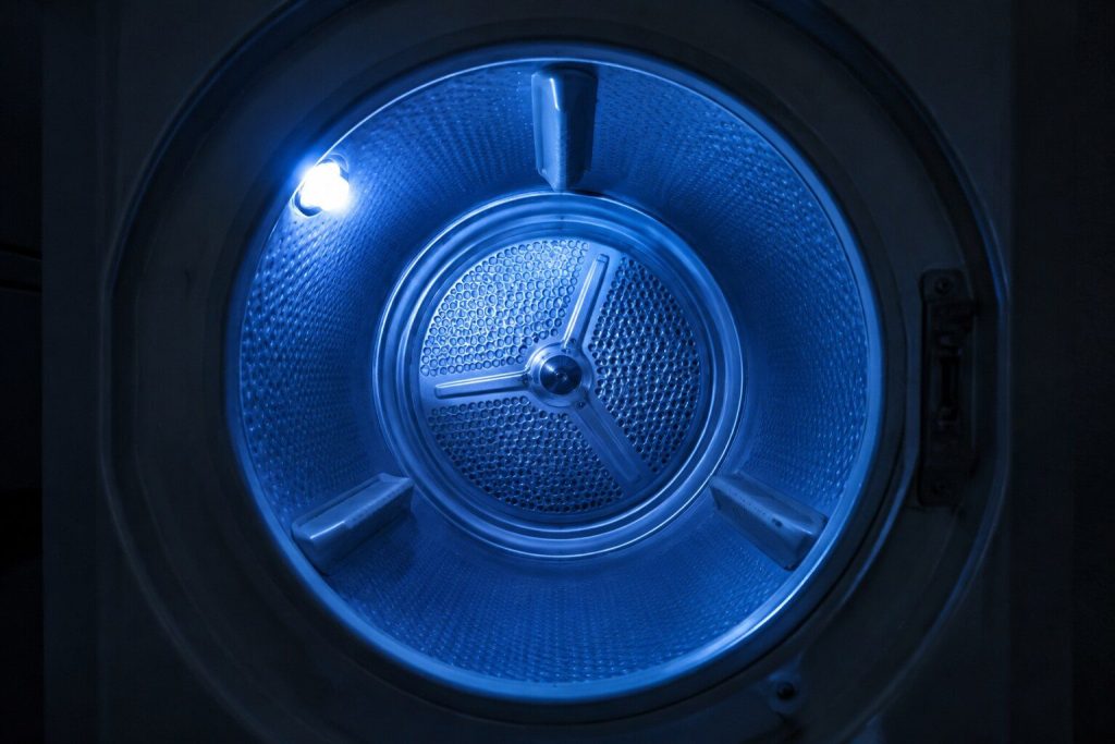 Looking straight into an empty dryer drum through the open door at the three molded lifting vanes standing off the curved drum wall, the perforated rear bulkhead a soft shape behind them.