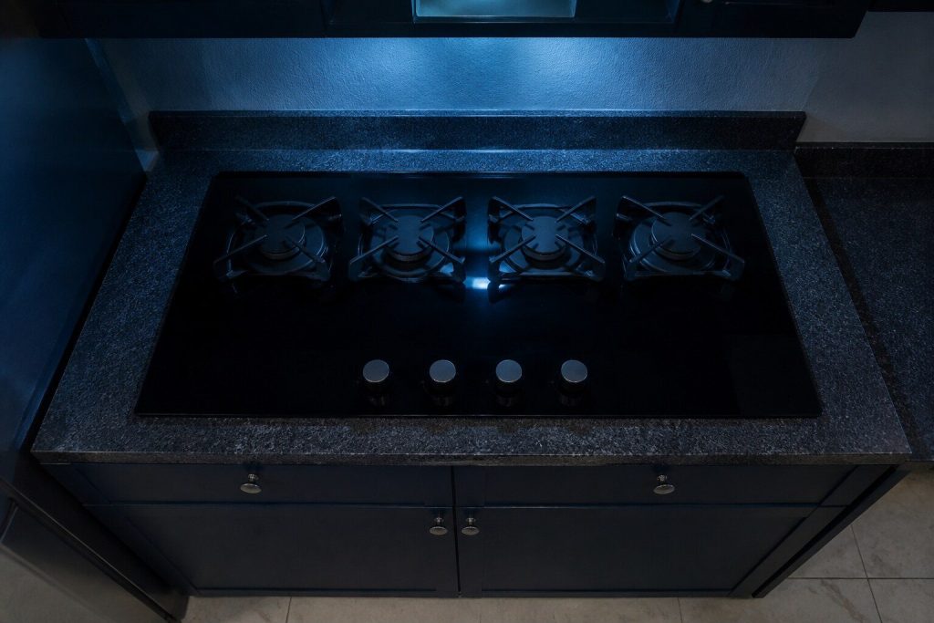 Four sealed gas burners standing on ONE unbroken sheet of black glass dropped into a counter, thin steel grates over them and a row of knobs along the front edge.
