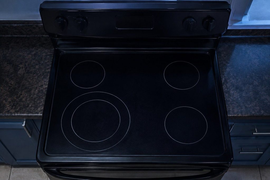 The smooth black ceramic top of a FREESTANDING electric range seen from directly above with its back console at the top of the frame: four printed element rings on the glass, the front left one a large double ring markedly wider than the other three.