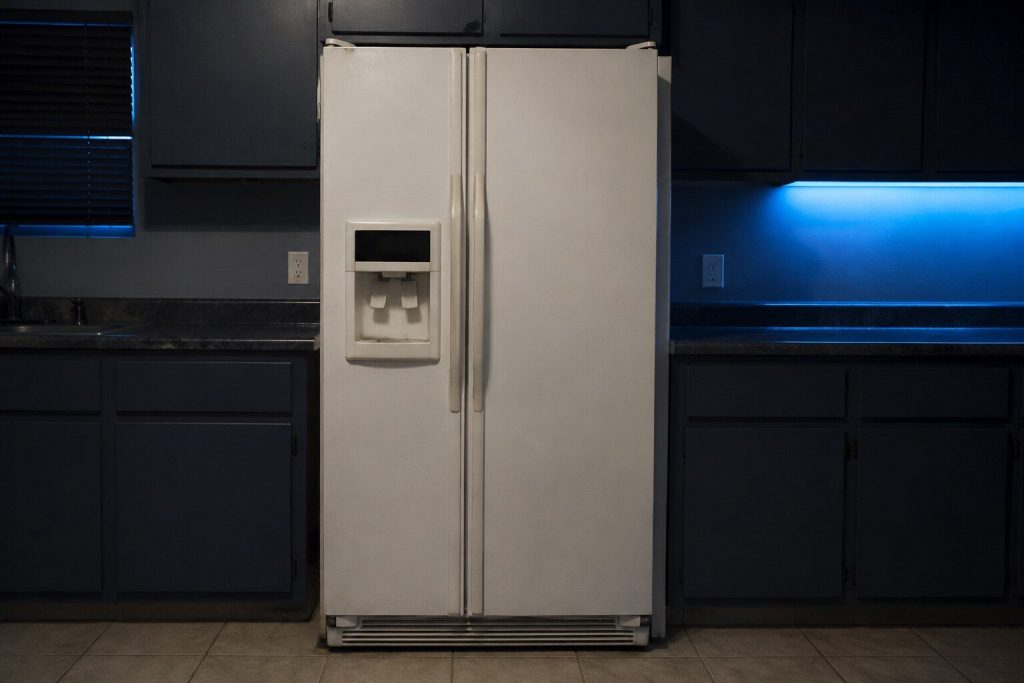 A white freestanding side-by-side refrigerator standing square between two runs of laminate base cabinets, both doors closed, a recessed ice and water dispenser set into the upper part of the narrow left door with a plain unmarked paddle and a dark unlit panel above it.