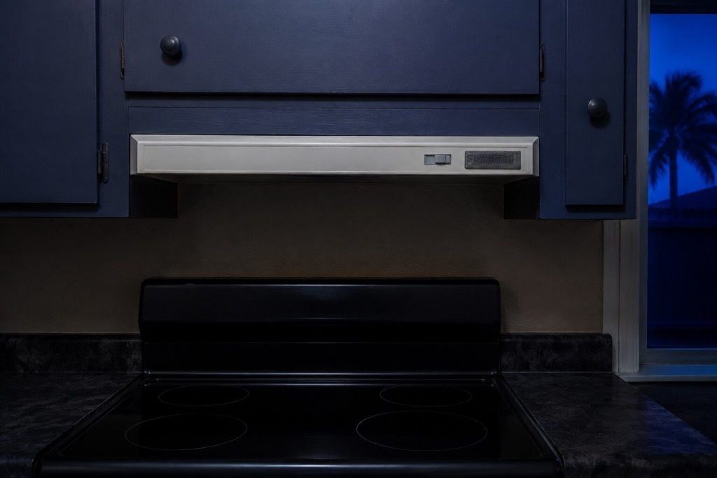 A flat rectangular under-cabinet range hood screwed to the underside of a wall cabinet directly above a cooktop, the cabinet door continuing above it and NO chimney or canopy of any kind.
