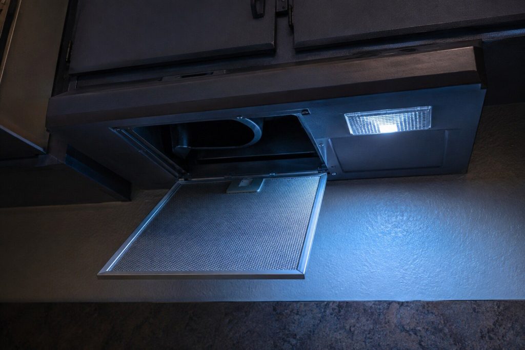 The underside of a flat under-cabinet range hood seen from below with one aluminum mesh grease filter unclipped and hanging down on its catch, the blower opening and the lamp lens visible in the recess above it.