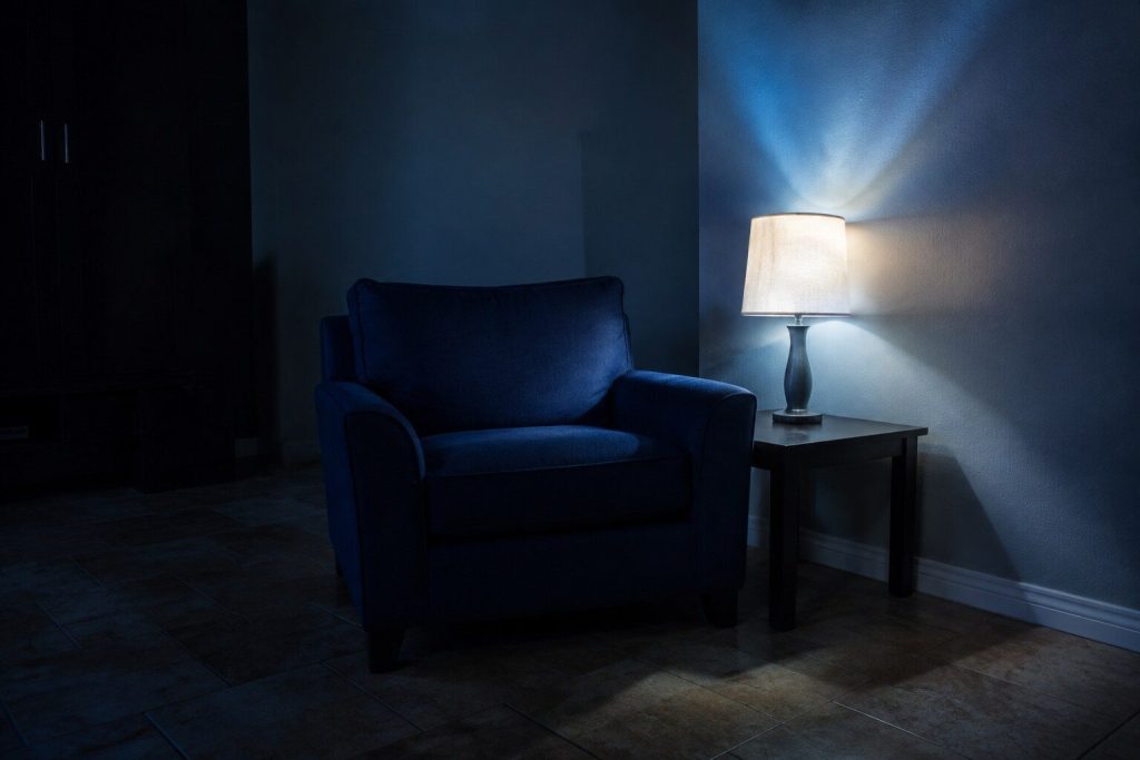 A corner of a living room at night: an armchair, a side table and one lit lamp, the rest of the room dark.