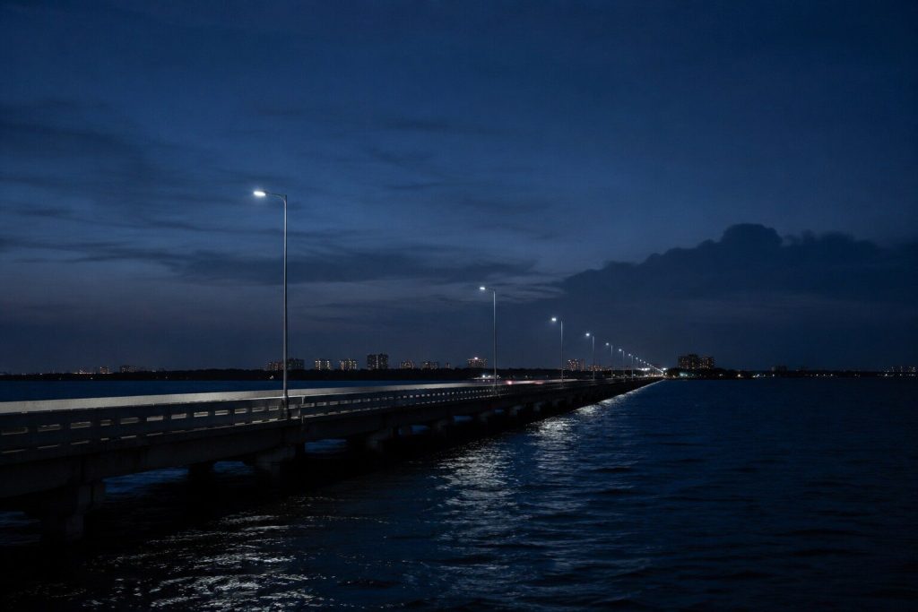 A causeway across dark water at dusk with a line of lamps running away into the distance and a low city skyline on the horizon.