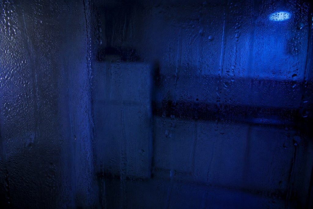 Condensation running in tracks down the inside of a dark kitchen window at night, the room behind it out of focus and blue.
