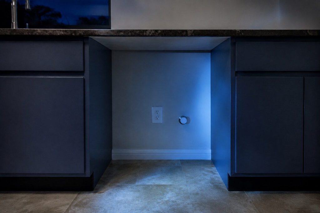 The narrow empty gap left between two runs of base cabinets where a machine stands, nothing in it: the vinyl floor running back to the wall, an outlet and a capped supply stub low at the back, the counter overhanging above.