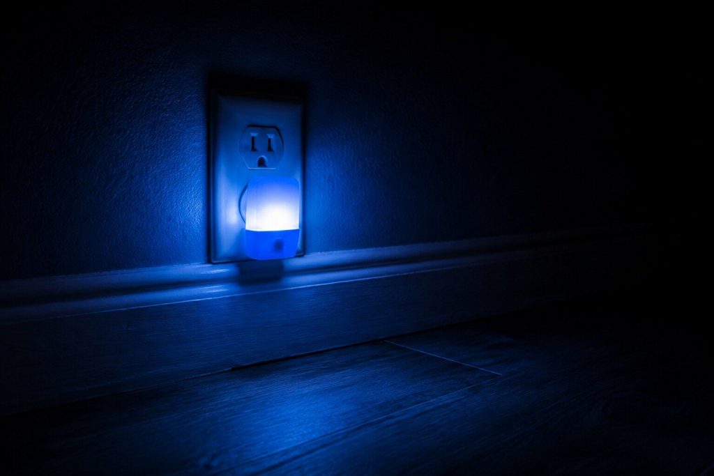 A small plug-in night light glowing in a baseboard outlet in an otherwise completely dark room, the skirting and a strip of vinyl floor lit for a hand's width around it and everything else black.