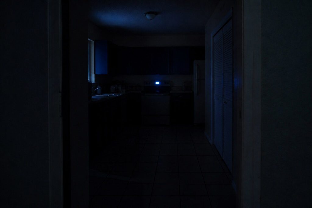 A dark kitchen at night seen wide from a doorway, one machine display glowing on the far side of the room as a soft unfocused shape of blue-white light and everything else in shadow.