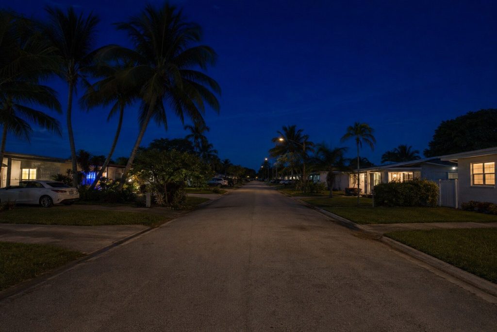 A South Florida residential street seen down its length at blue hour, low houses and palms either side, warm windows just coming on and the sky deep navy above.