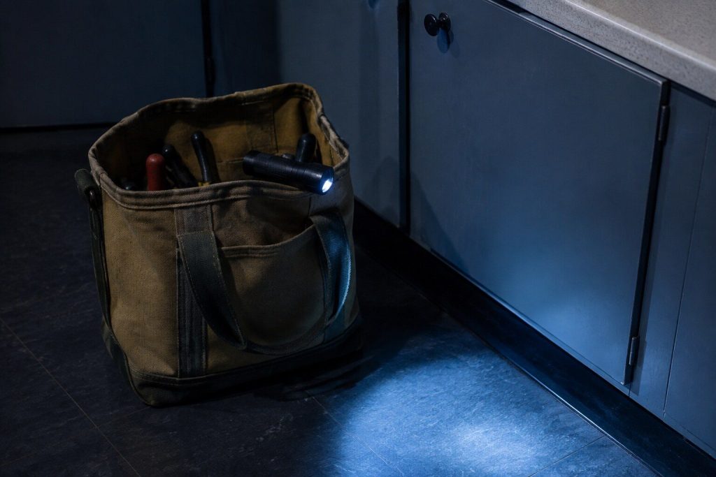 An open canvas tool bag standing closed-up on a dark vinyl tile floor beside a kickboard, a torch lying on top of it throwing a low pool of light across the tile.