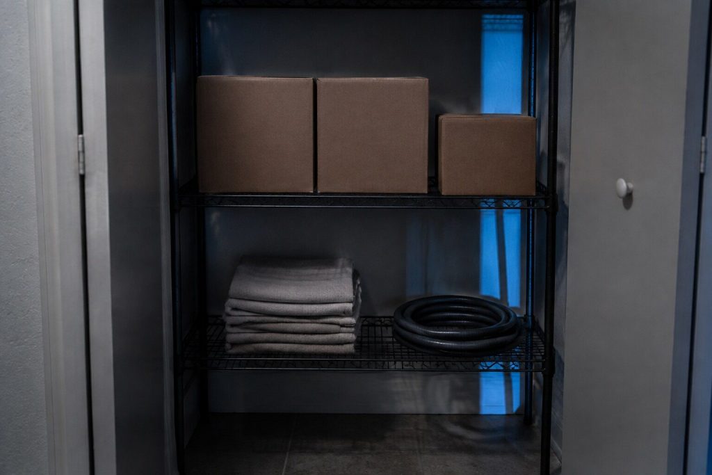 A wire utility shelf in a dim closet holding plain unlabeled cardboard cartons, a folded stack of cloths and a coil of hose.