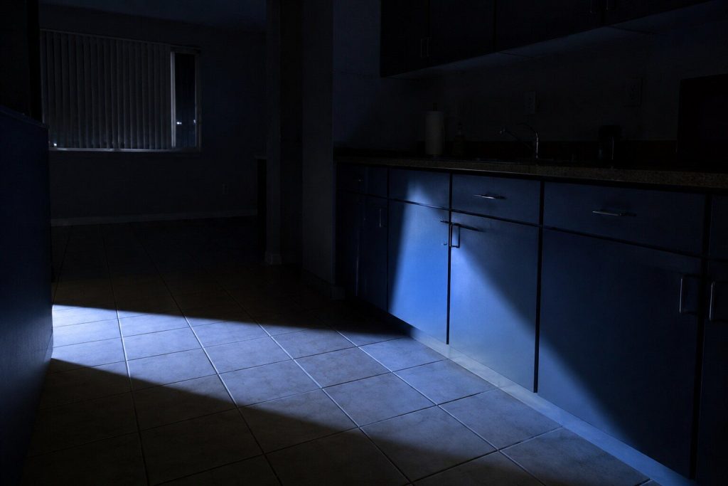 A kitchen at night lit only by one open machine door out of shot, its interior lamp throwing a hard bright wedge of light across a dark tile floor and up the cabinet fronts opposite.