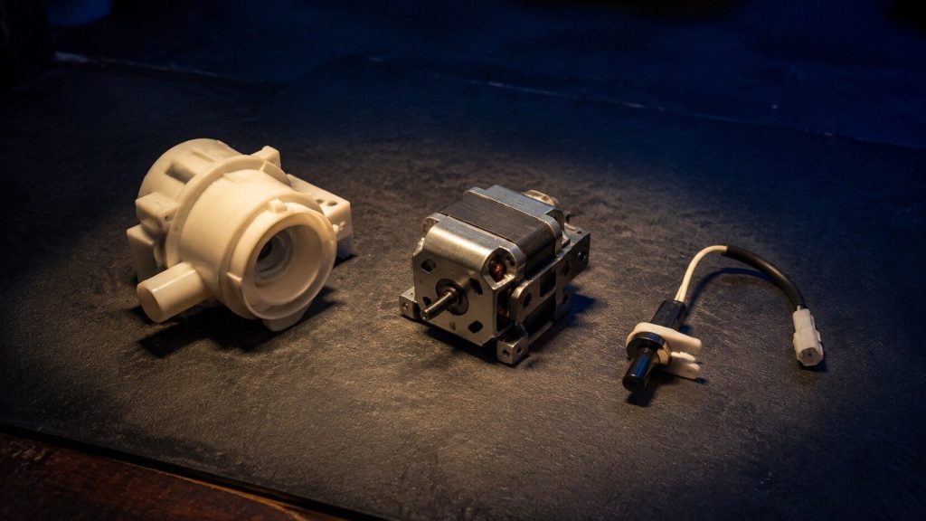 "Three small unbranded appliance components laid out on a clean workbench mat under a single work light — a pump housing, a small motor and a sensor with a lead" — the parts that get blamed on the board, which is when the page says the board is NOT the answer