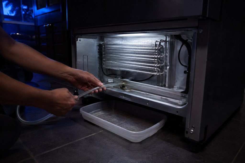"A length of flexible tubing being fed into the drain trough at the bottom of an evaporator compartment ... a shallow tray under the machine" — the blocked defrost drain, which is the first of the four sources this page lists