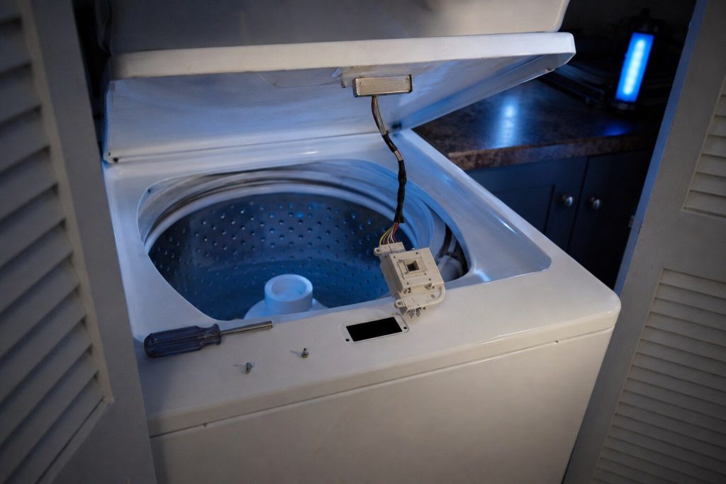 The lid lock housing lifted out of its cutout in the rim of a white top-load washing machine and hanging on its wiring loom over the open tub, the metal striker on the underside of the raised lid above it, a nut driver and two screws lying on the machine's top panel.