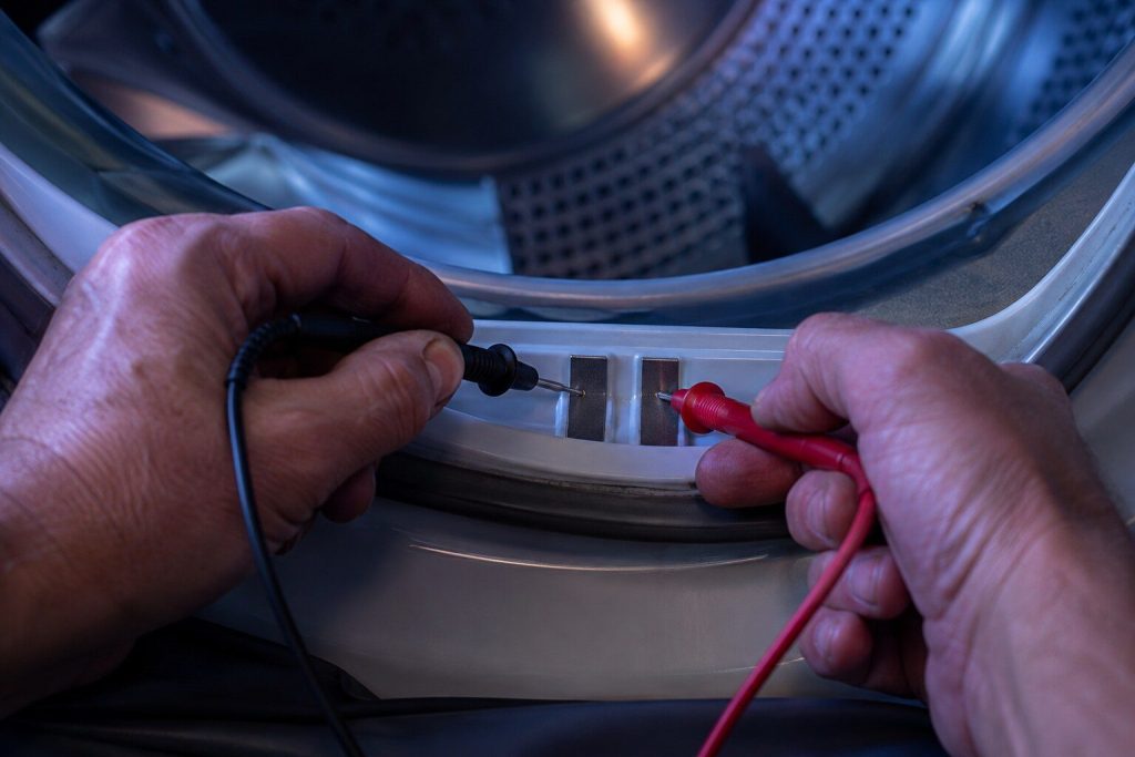 "Two meter probes held against the pair of metal sensor strips just inside the front of a dryer drum" — the two strips the automatic cycle reads, which is the whole subject