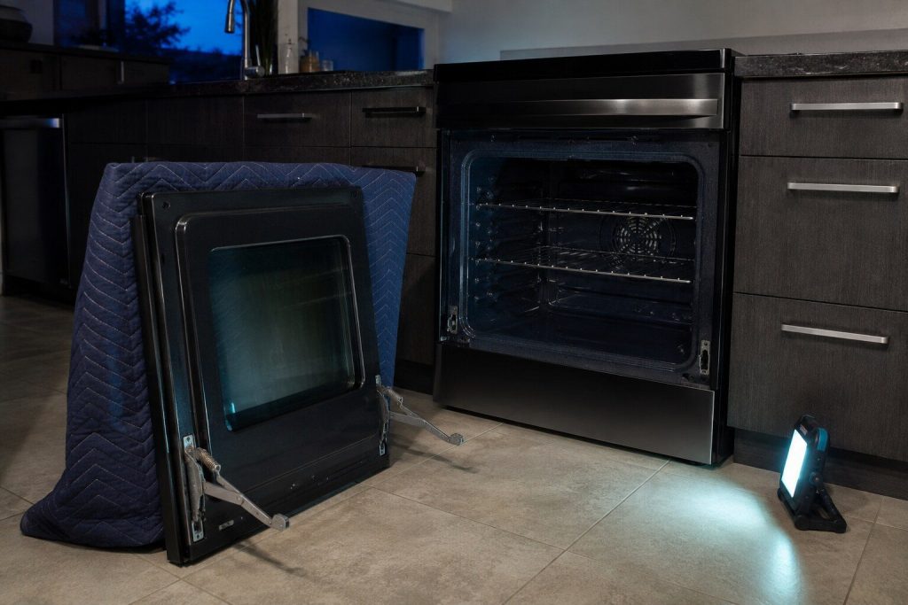 "An oven door lifted off the range and stood upright against a blanket on the floor, both hinge arms locked open, the empty hinge slots in the frame behind it"
