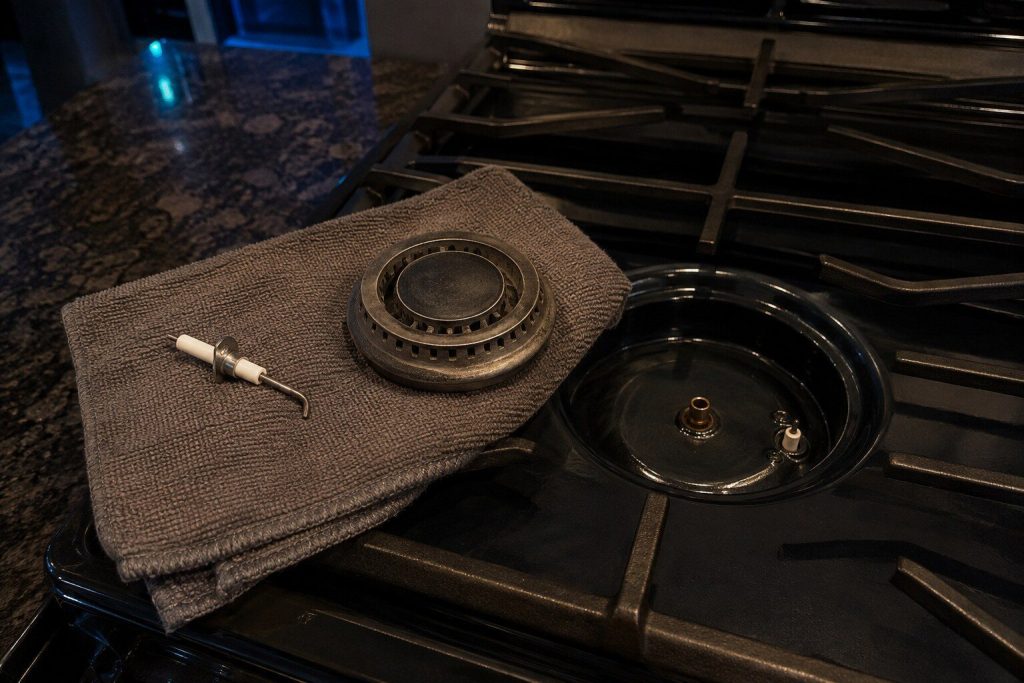 "A spark igniter and a burner head lying apart on a folded towel on a cooktop, the empty burner base and its orifice visible beside them" — the endless click is one of these two