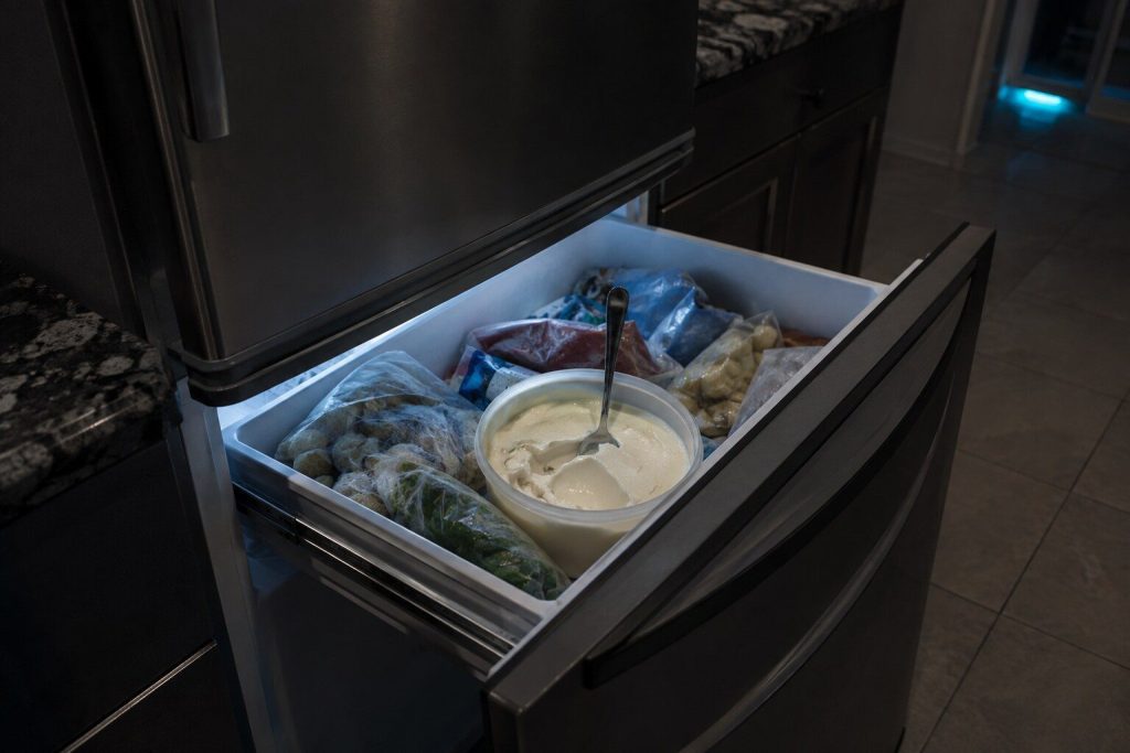 "An open tub of ice cream gone soft and glossy in a freezer drawer, a spoon standing upright in it, the packets around it slumped rather than rigid" — ice cream soft, which is the caption word for word