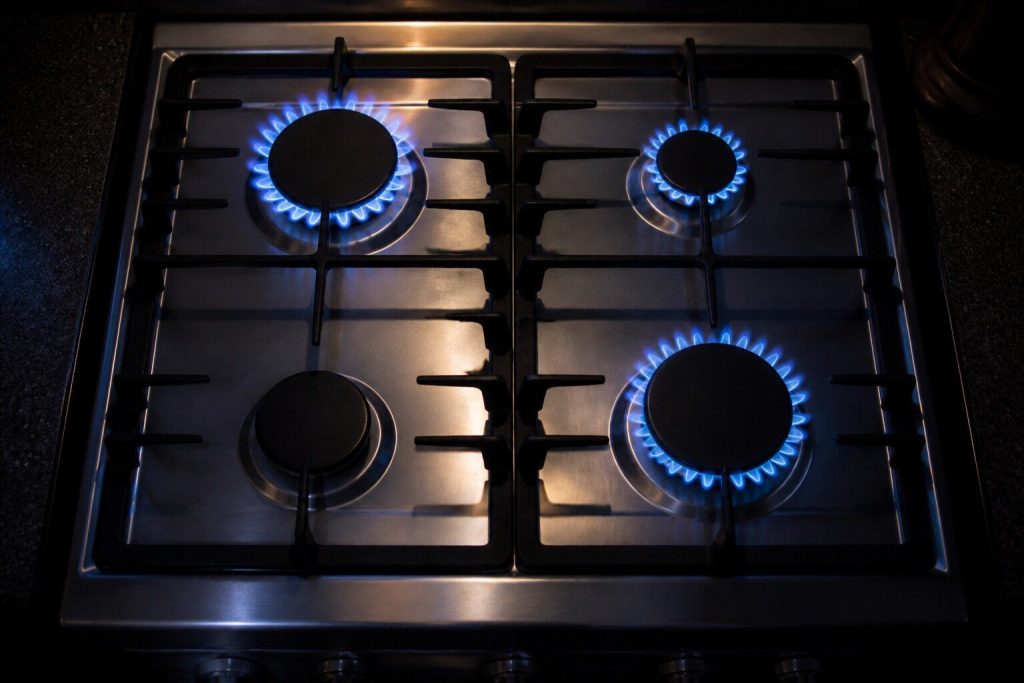 "A gas cooktop photographed from above at night with three burner crowns carrying steady blue flame and the fourth completely dark and cold" — the clicking burner, with the working ones beside it for contrast