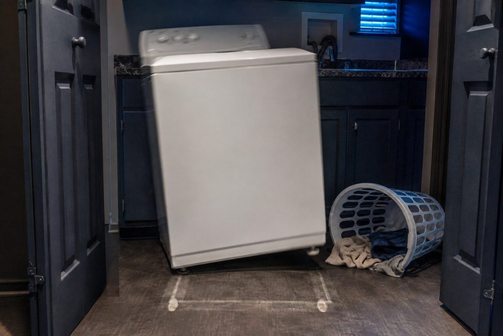 A white top-load washing machine part-way through a spin in a laundry closet at night, shifted forward out of the clean rectangle its feet had worn into the vinyl floor, one front foot lifted clear of the floor and the cabinet leaning on the other three, a plastic laundry basket knocked over beside it.